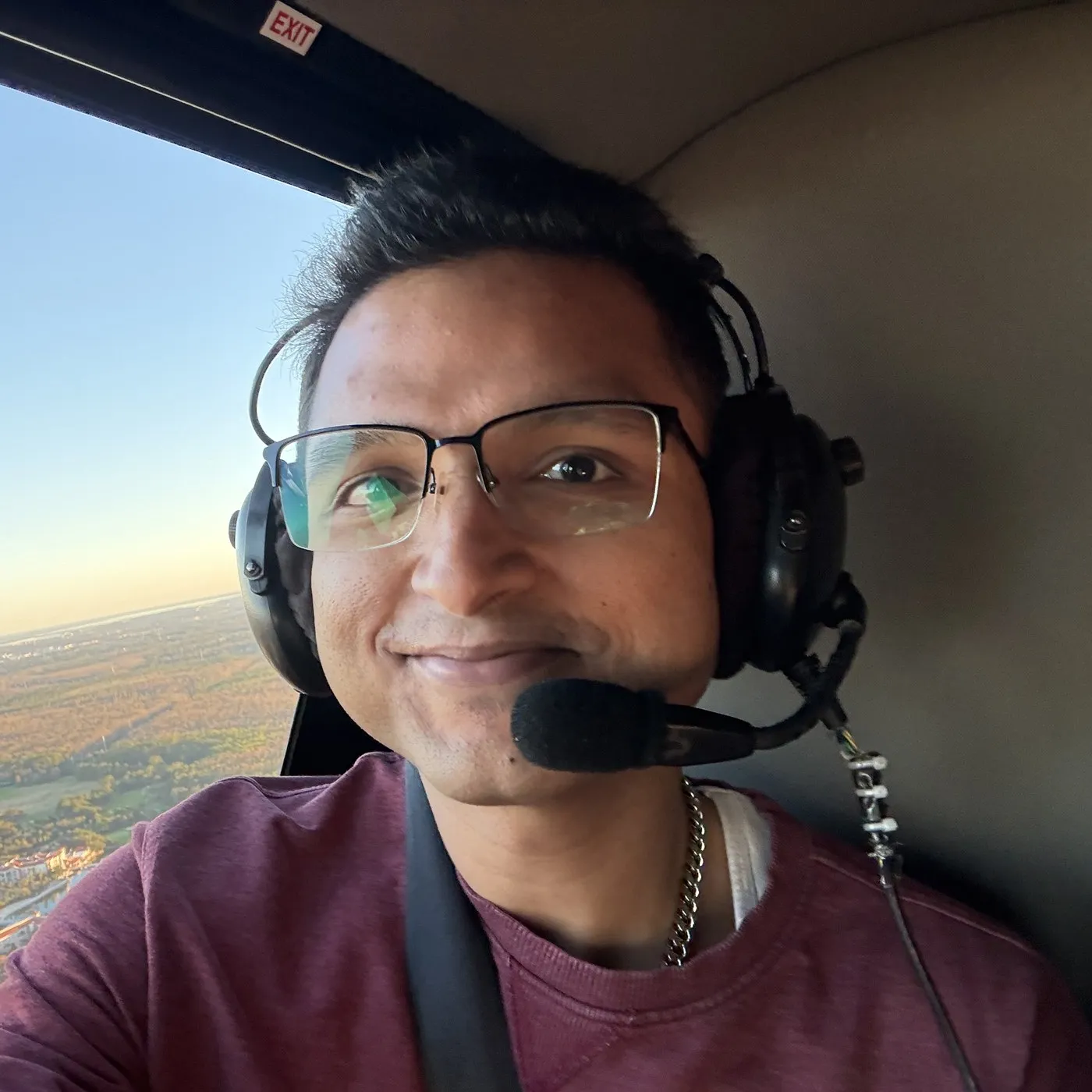 Kshitij Anand in the cockpit of a light aircraft, wearing an aviation headset, with farmland and a river far below.